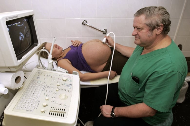 A very pregnant woman is laying on her back on a table with her huge stomach exposed as an older man in green scrubs performs an ultrasound on her.