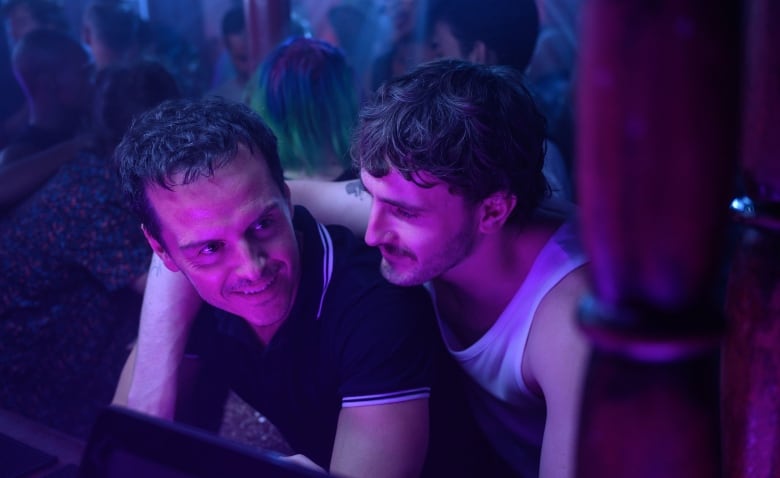 Still frame from the film All Of Us Strangers. Paul Mescal drapes an arm around Andrew Scott, cast in purple and blue lighting.