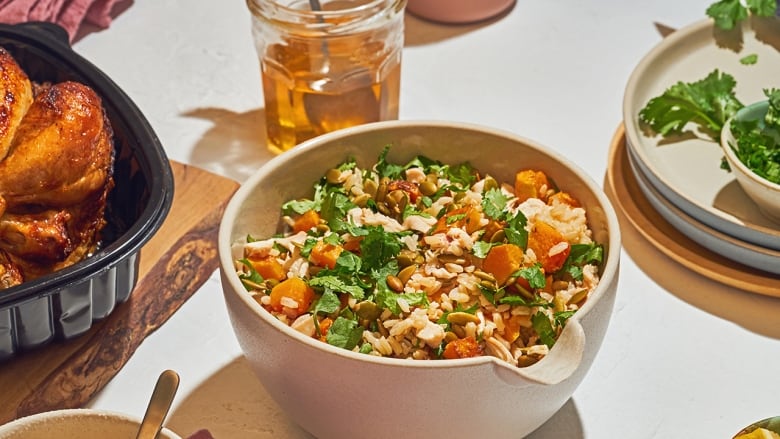 A beige bowl filled with Brown Rice, Chicken and Butternut Squash Salad on a beige surface. A rotisserie chicken sits on the left of the image and a stack on plates on the right. 