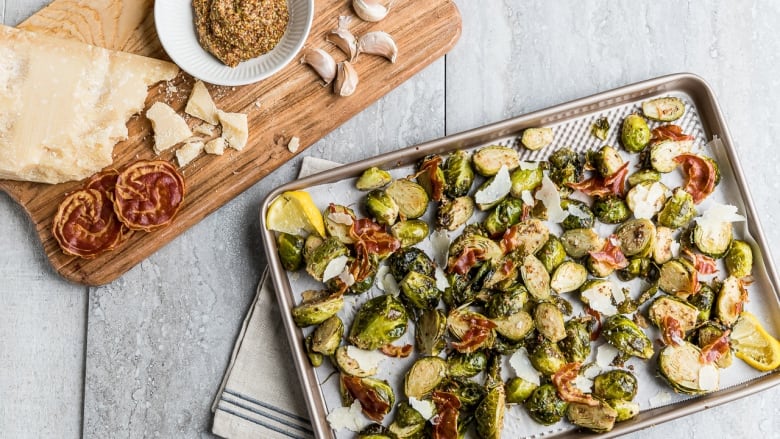 A baking sheet with roasted Brussels sprouts and pancetta on it. A wooden cutting board with grainy mustard, a block of parmesan cheese and pancetta sit next to the baking sheet. 