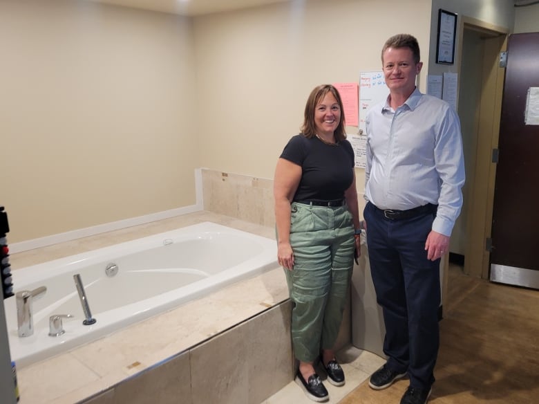 Two smiling people stand in a room next to an empty bathtub.