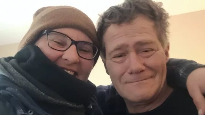 Two people pose for a selfie, on the left a woman in a toque and a scarf smiles, on the right a grey-haired man smiles into the camera. 