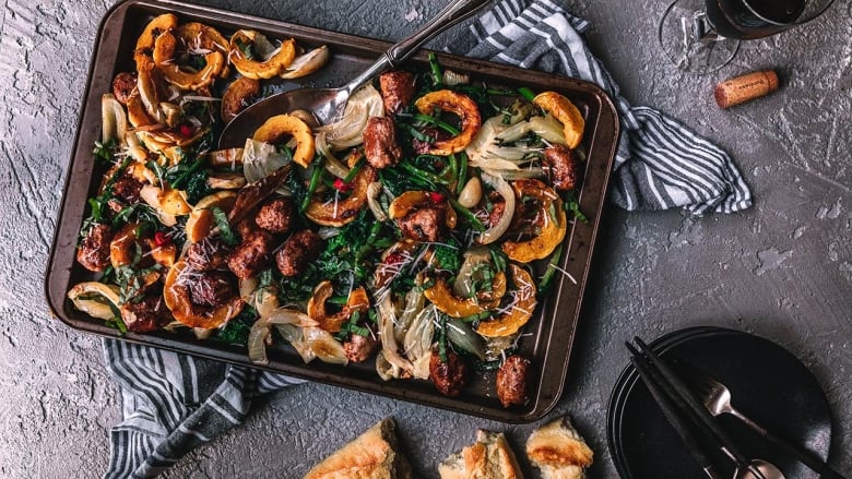 Overhead shot of a sheet pan on a dark grey surface. Slices of roasted delicata squash, onion, pieces of sausage and roasted rapini are on the pan. 