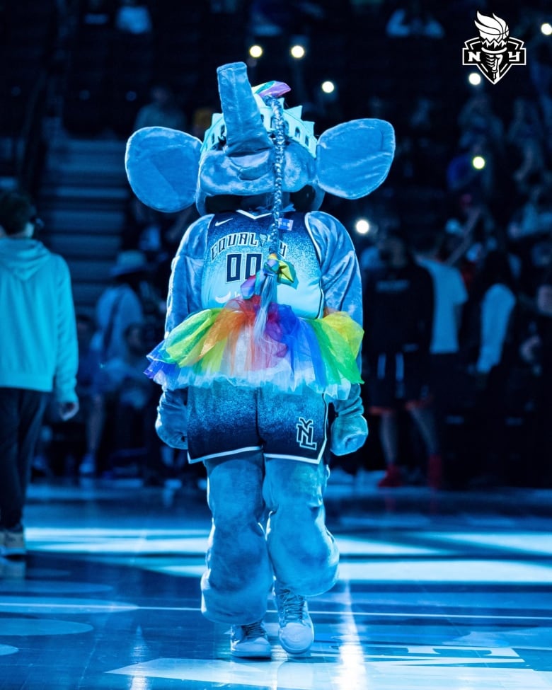 A person in an elephant costume stands in the middle of a basketball court, head titled back and lit in bright blue. She's sporting a basketball uniform and a rainbow tutu. A single long braid is strewn across her elephant face.