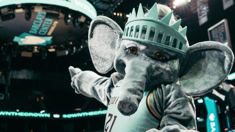 Close up of a person on an elephant costume, sporting a basketball uniform and a crown like the Statue of Liberty's, and pointing to a sign on a stadium ceiling that reads 'Barclays Center'
