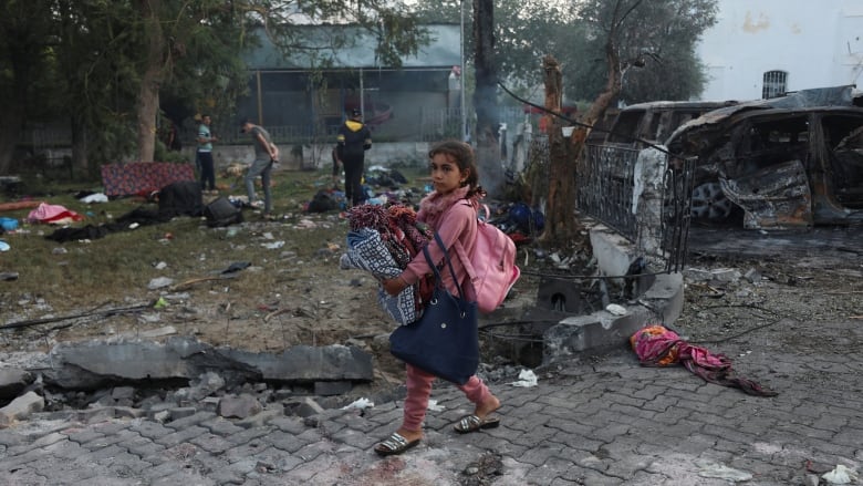 A young girl wearing a pink shirt and pink backpack carries her belongings past the ruins of a community hospital.