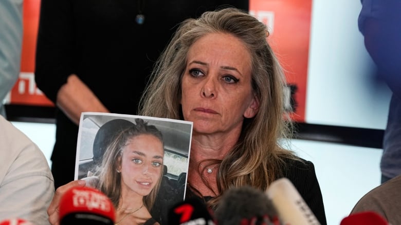 A woman who looks distressed holds up a picture of another, younger woman while seated in front of microphones.