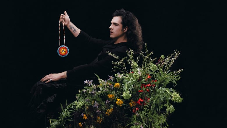 Side profile of musician Jeremy Dutcher holding a beaded necklace, surrounded by flowers. 