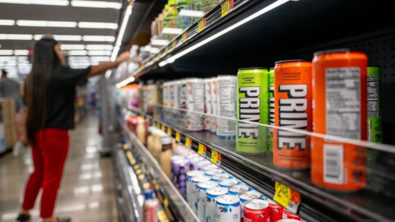 Brightly coloured cans with the word "PRIME" in white capital letters are seen on store shelves. A woman is visible in the background reaching for a shelf.