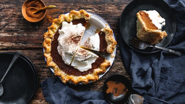 Overhead shot of a pumpkin pie topped with whipped cream on a wooden table. A slice of the pie is sitting on a black plate. 