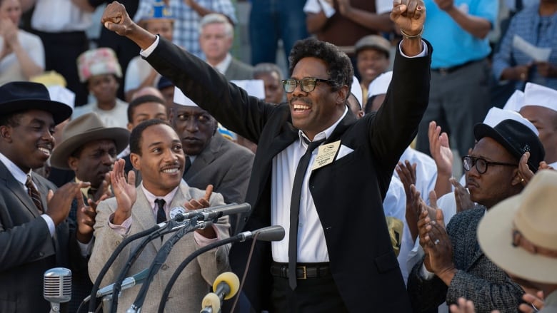 Still frame from the film Rustin. Colman Domingo raises his arms and grins widely, standing in front of microphones at a podium, with an audience of fellow Black people clapping behind him.