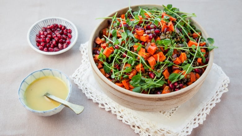 A bowl of lentil salad with chunks of sweet potatoes in it, and topped with microgreens and pomegranate arils. Small bowls of pomegranate arils and salad dressing sit next to the salad. 
