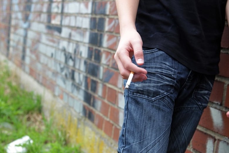 A young person stands next to a brick wall holding a cigarette.