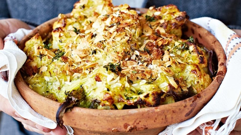 Closeup on a baking dish with cheesy, baked cauliflower and broccoli. Two hands are holding the dish with a white kitchen towel. 